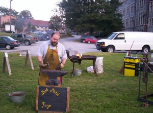 Geof hammering metal at the anvil. In the background the coal fire smokes merrily.