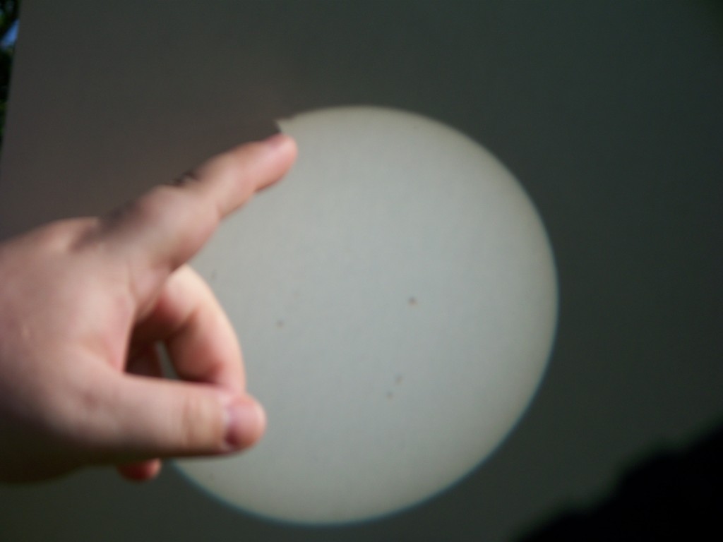 The projected image of the sun on white posterboard. A finger points at the shadow of Venus as it first touches the sun.