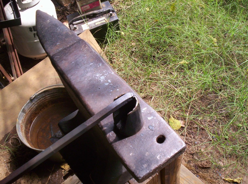 The metal stock has been cut off and one end rests on the hardie tool that is resting on the anvil.