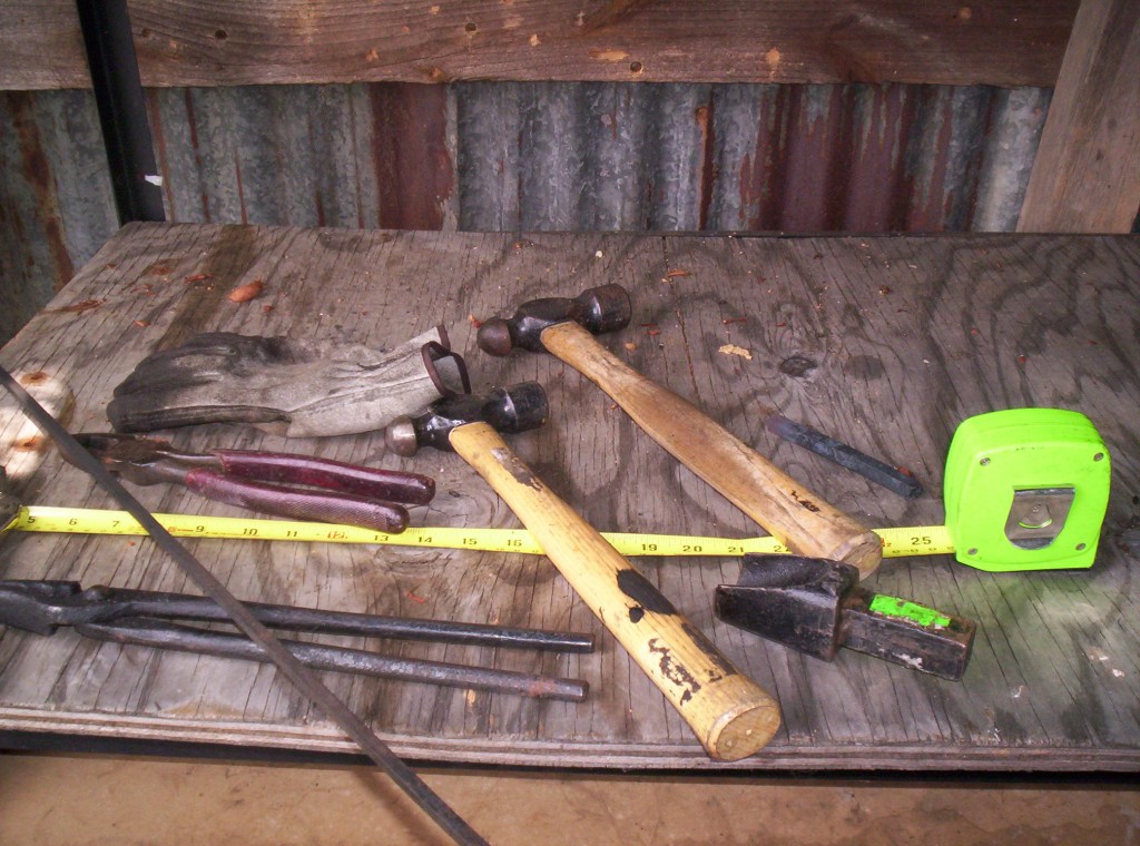 A pile of tools on the workbench, two hammers, tongs, pliers, hardie tool, tape measure and metal stock.