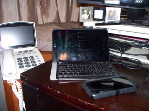A cellphone, tablet with keyboard and caption phone on a desk.