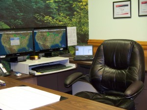 A picture of Geof's desk. Multi monitors, a netbook and desk blotter surround an empty chair.